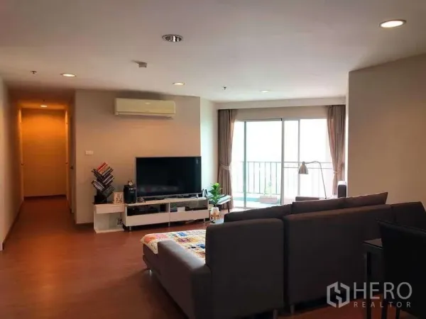 Condominium for sale in Huai Khwang, Bangkok - Spacious living room with sectional sofa, TV console, wood floors and sliding doors to a bright balcony.