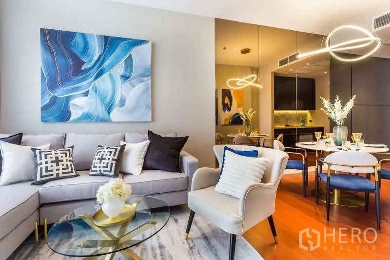 公寓 出租在Watthana, Bangkok - 时尚客厅，转角沙发、扶手椅、玻璃茶几与蓝色装饰画。
