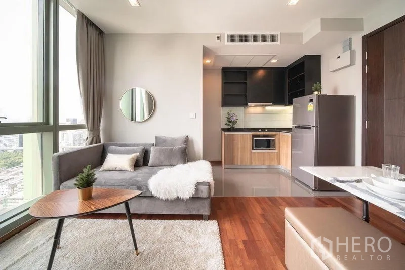 Condominium for rent in Ratchathewi, Bangkok - Open-plan living with gray sofa, round mirror, and kitchen in the background on polished floors.