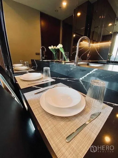 Condominium for rent in Khlong Toei, Bangkok - Breakfast bar with black stone countertop, sink, and place settings for two.