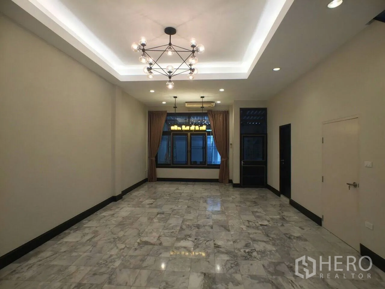 Townhouse for rent in Khlong Toei, Bangkok - Spacious living room with marble flooring, recessed ceiling and chandelier, and curtained windows.