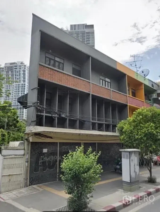 店屋 出租在Khlong Toei, Bangkok - 现代风格多层店屋外观，灰色与红砖立面，转角位置，一层卷闸门，周边有行道树和高层公寓，位于曼谷Ekkamai。