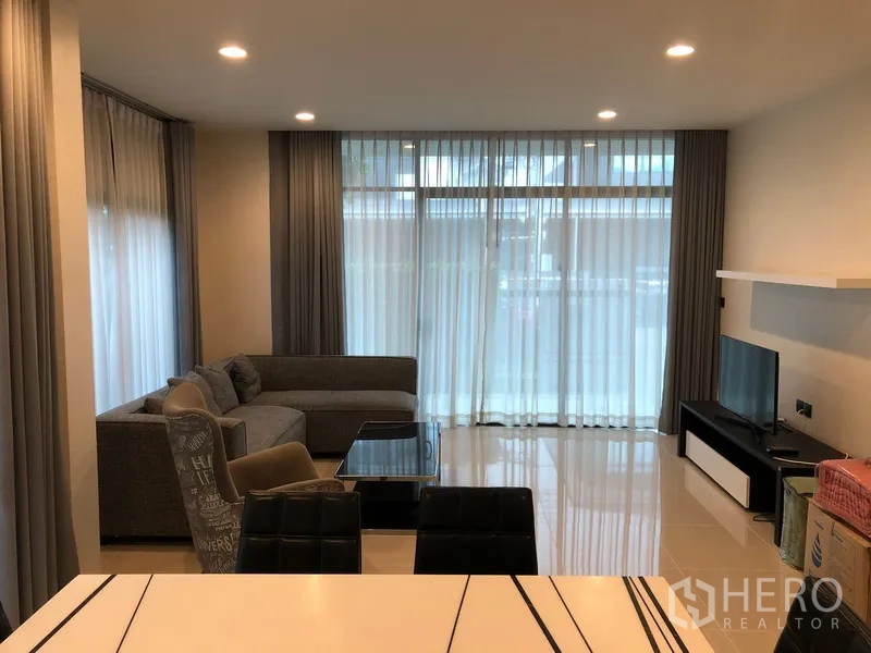 Detached House for rent or sale in Bang Kapi, Bangkok - Bright living room with L-shaped sofa, TV console, recessed lights and full-height sheer curtains.