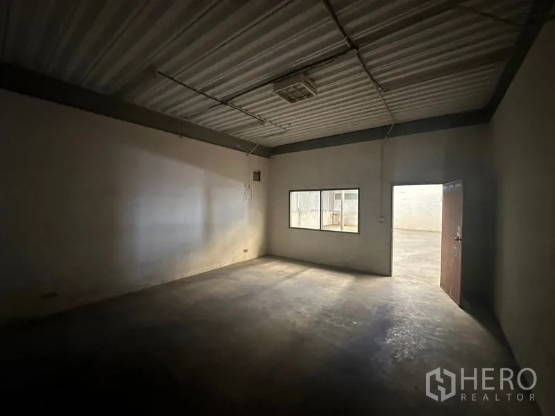 Warehouse for rent in Suan Luang, Bangkok - Office room with window and door opening to the main warehouse floor, exposed lighting above.