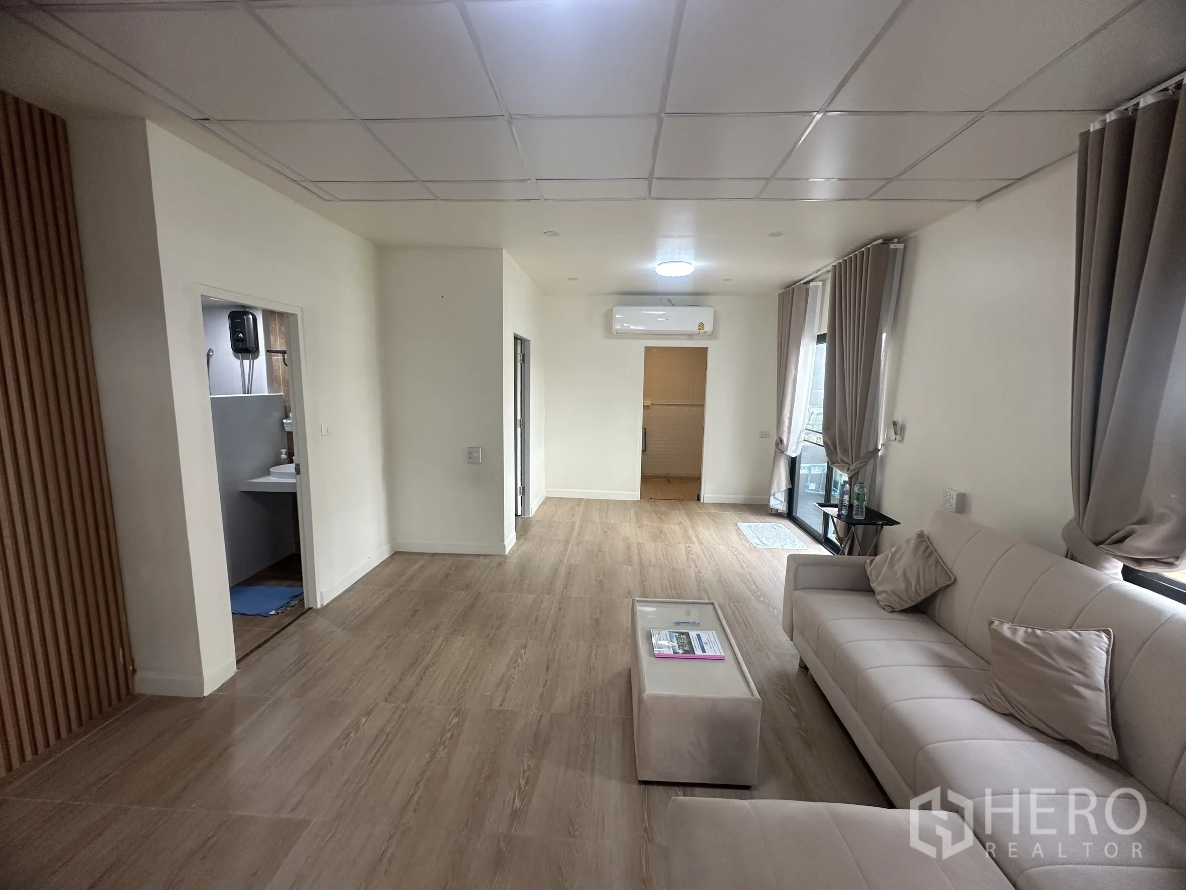 联排别墅 for sale in Lam Luk Ka, Pathum Thani - 明亮客厅，设有沙发、窗帘、木纹地砖与空调。