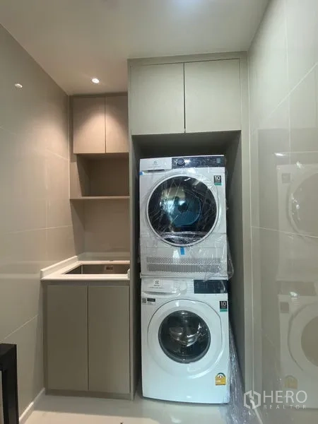 Condominium for rent in Watthana, Bangkok - Compact laundry room with stacked washer and dryer, sink, and storage cabinets.