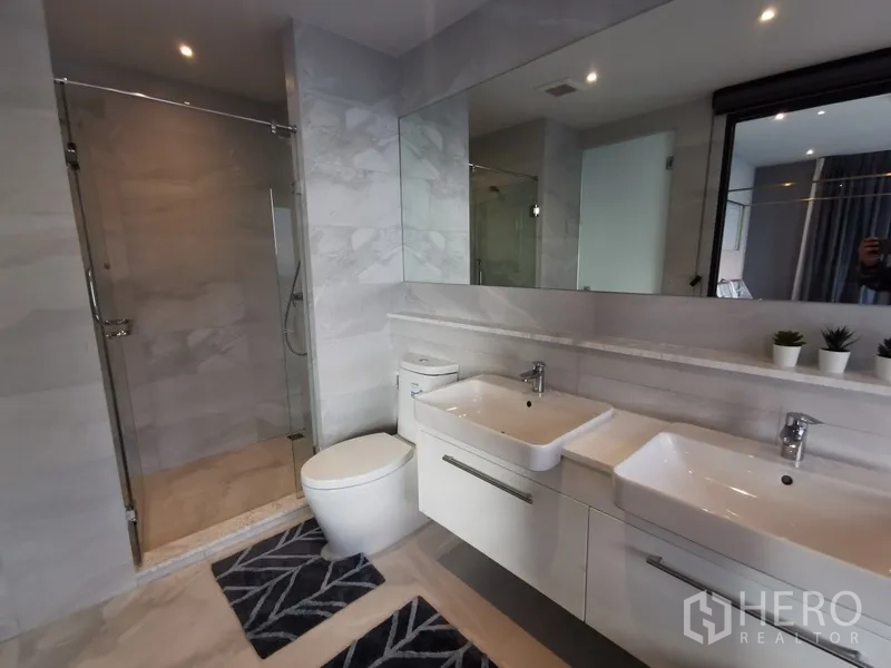 Condominium for rent or sale in Watthana, Bangkok - Contemporary bathroom with glass-enclosed shower, twin sinks, wide mirror, and neutral stone tiles.