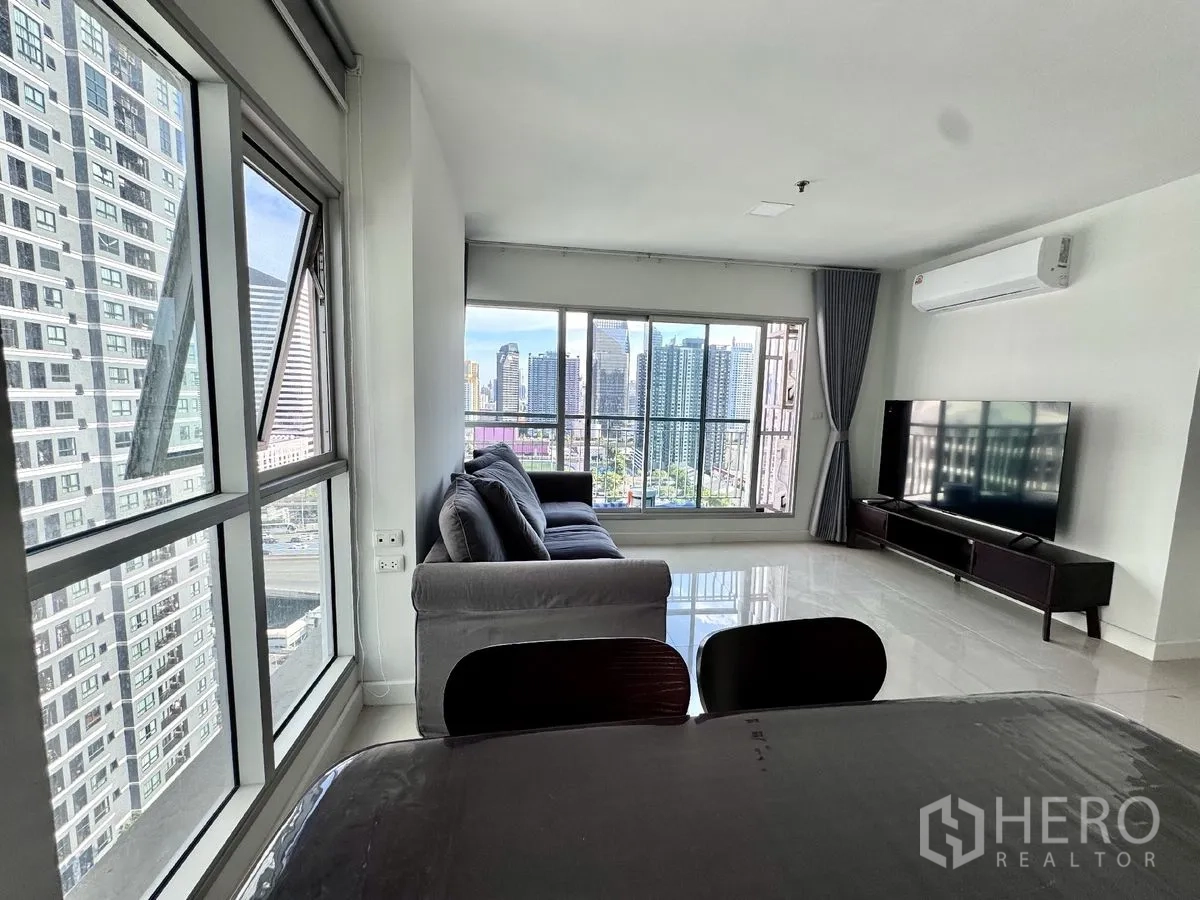 Condominium for rent in Huai Khwang, Bangkok - Bright living room with large windows, gray sofa, TV console, and a city view.