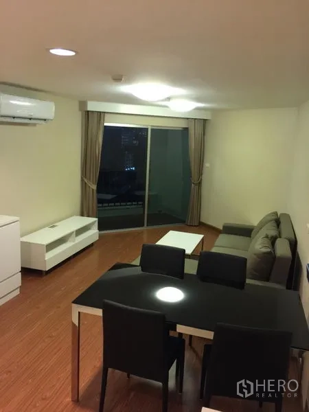 Condominium for rent in Huai Khwang, Bangkok - Living room with gray sofa, dining set and sliding glass door to a balcony at night.