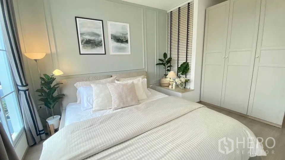Townhouse for rent in Prawet, Bangkok - Serene bedroom with panelled feature wall, crisp white bedding, floor lamp, built-in wardrobe and potted plants.