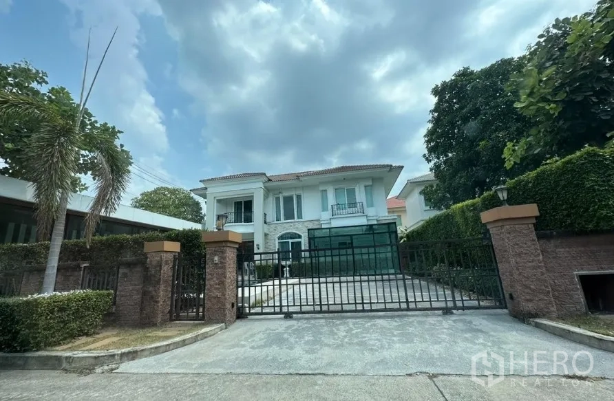 Detached House for sale - Front view of brick-fenced residence with sliding iron gate, hedge-lined boundary and mature trees under a cloudy sky.