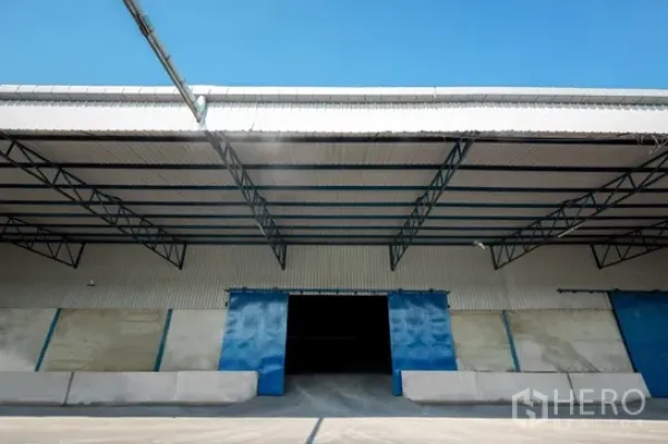 货栈 出租在Pathum Thani - 仓库外立面，钢结构雨棚下设有装卸平台和蓝色推拉门。