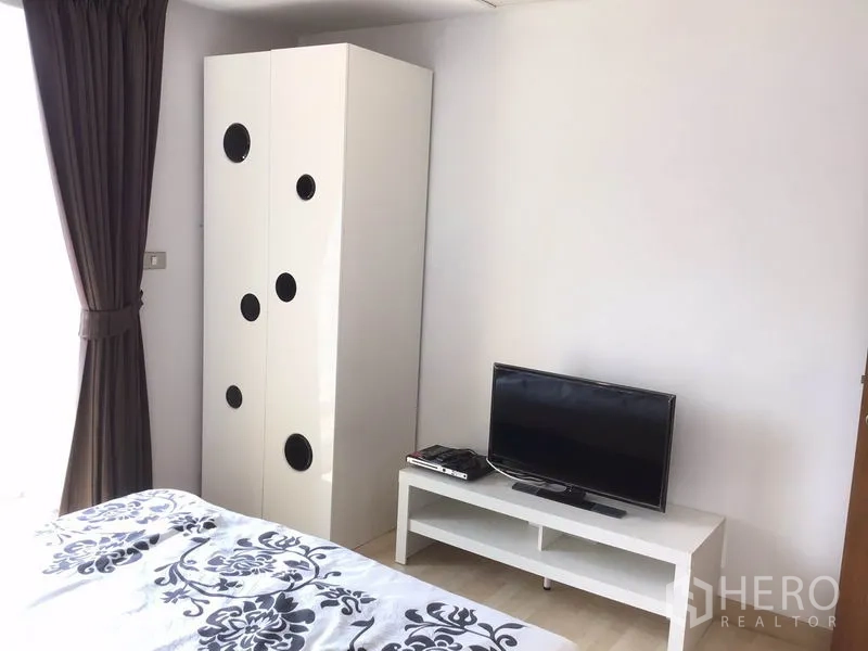 公寓 出租在Watthana, Bangkok - 卧室一角，落地窗帘，高柜带圆形装饰孔，低矮电视柜上摆放电视。