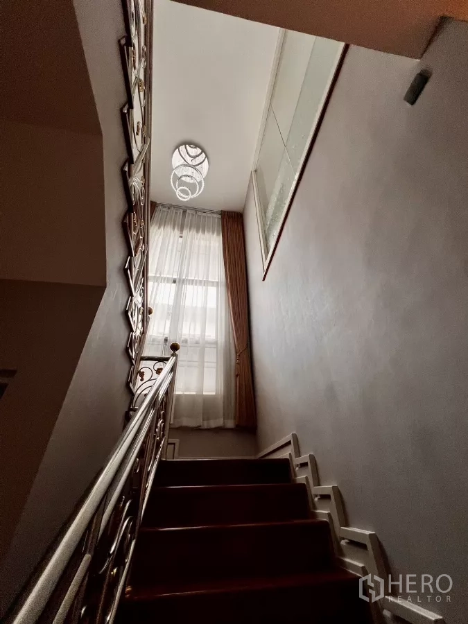 Detached House for rent or sale in Prawet, Bangkok - Staircase with decorative metal handrail and chandelier beside a tall window.