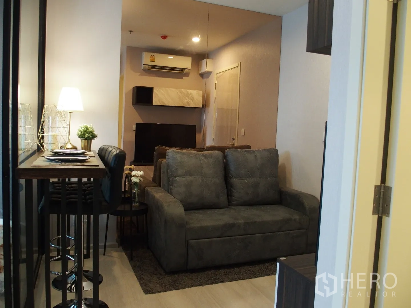Condominium for rent in Huai Khwang, Bangkok - Cozy living room featuring a gray sofa, high-top dining set, built-in shelves, and air conditioning.