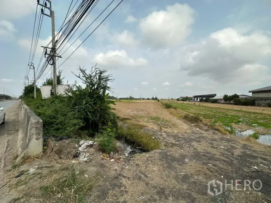 Land for sale in Bang Bua Thong, Nonthaburi - Corner of the plot showing dry soil, canal edge and greenery next to the main road and utilities.