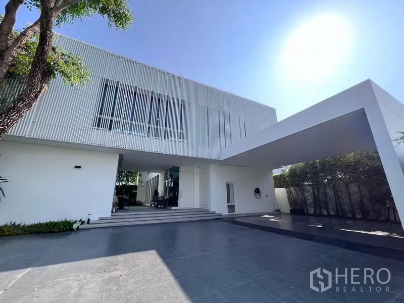 Detached House for sale in Samut Prakan - Front driveway and covered parking of the modern white house under a bright sky.
