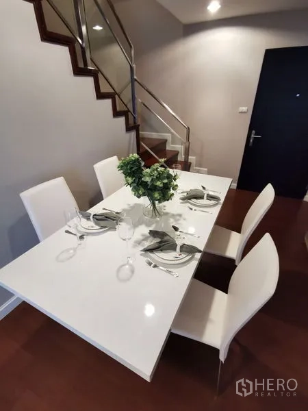 Condominium for rent or sale in Huai Khwang, Bangkok - Contemporary dining area with a white table and four chairs beside a wooden staircase.