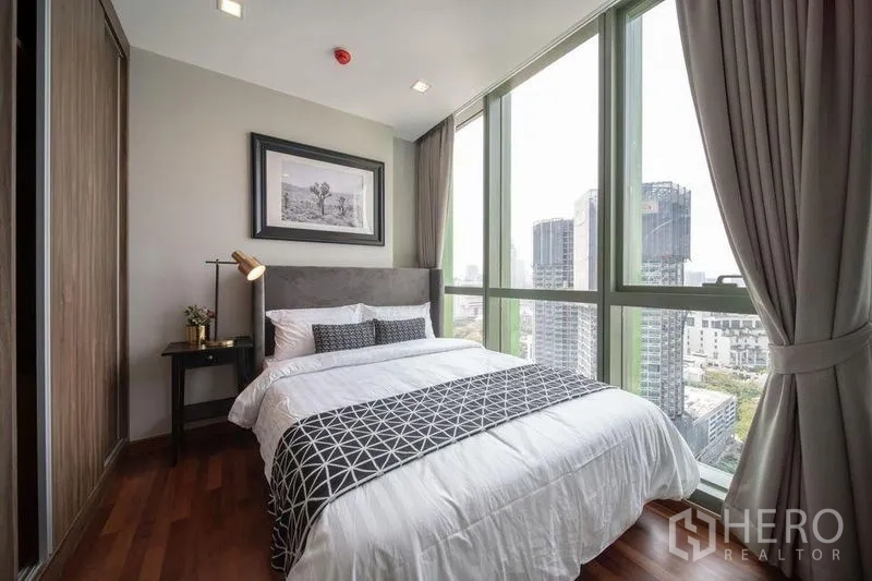 Condominium for rent in Ratchathewi, Bangkok - Corner bedroom with floor-to-ceiling glass, city skyline view, and a double bed with grey headboard.