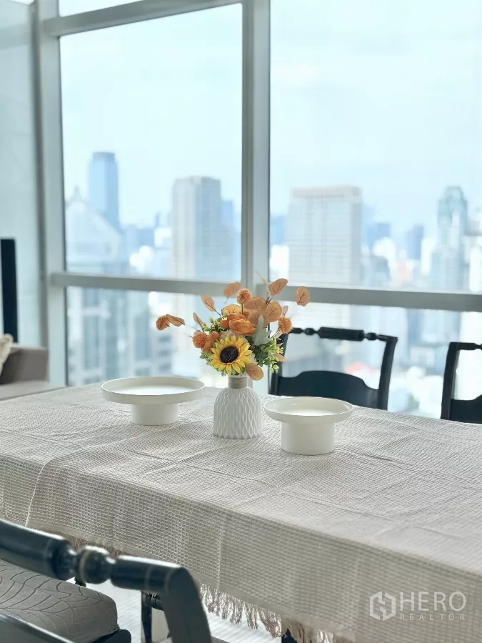 คอนโด ให้เช่าในPathum Wan, Bangkok - โต๊ะอาหารพร้อมแจกันดอกไม้ ตั้งอยู่หน้าหน้าต่างกระจกกว้างมองเห็นวิวเมือง