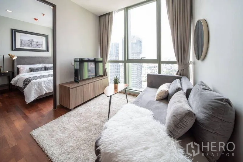 Condominium for rent in Ratchathewi, Bangkok - Sunlit lounge with plush sofa, rug, TV stand and bedroom entry beside tall glass windows.