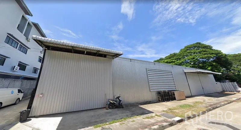 Warehouse for rent in Wang Thonglang, Bangkok - Exterior of a corrugated metal warehouse with wide sliding doors and driveway access beside trees under a clear sky.