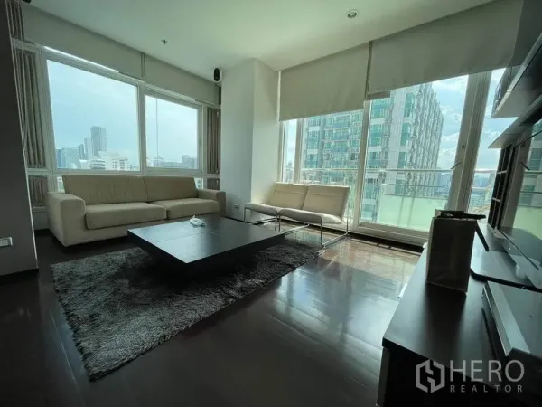 Condominium for rent or sale in Watthana, Bangkok - Bright living room with floor-to-ceiling windows, beige sofas, dark wood floors and city skyline views.