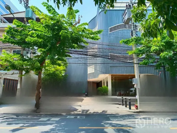 独立商业地产 出租在Bang Rak, Bangkok - 街景正面，灰色建筑一层玻璃立面与弧形阳台，临街人行道绿荫浓密，靠近轻轨出入口。