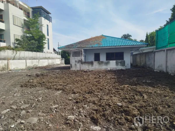 Detached House for rent or sale in Bang Na, Bangkok - Rear view of the detached house with blue roof and a large cleared soil plot bordered by walls and nearby mid-rise buildings.