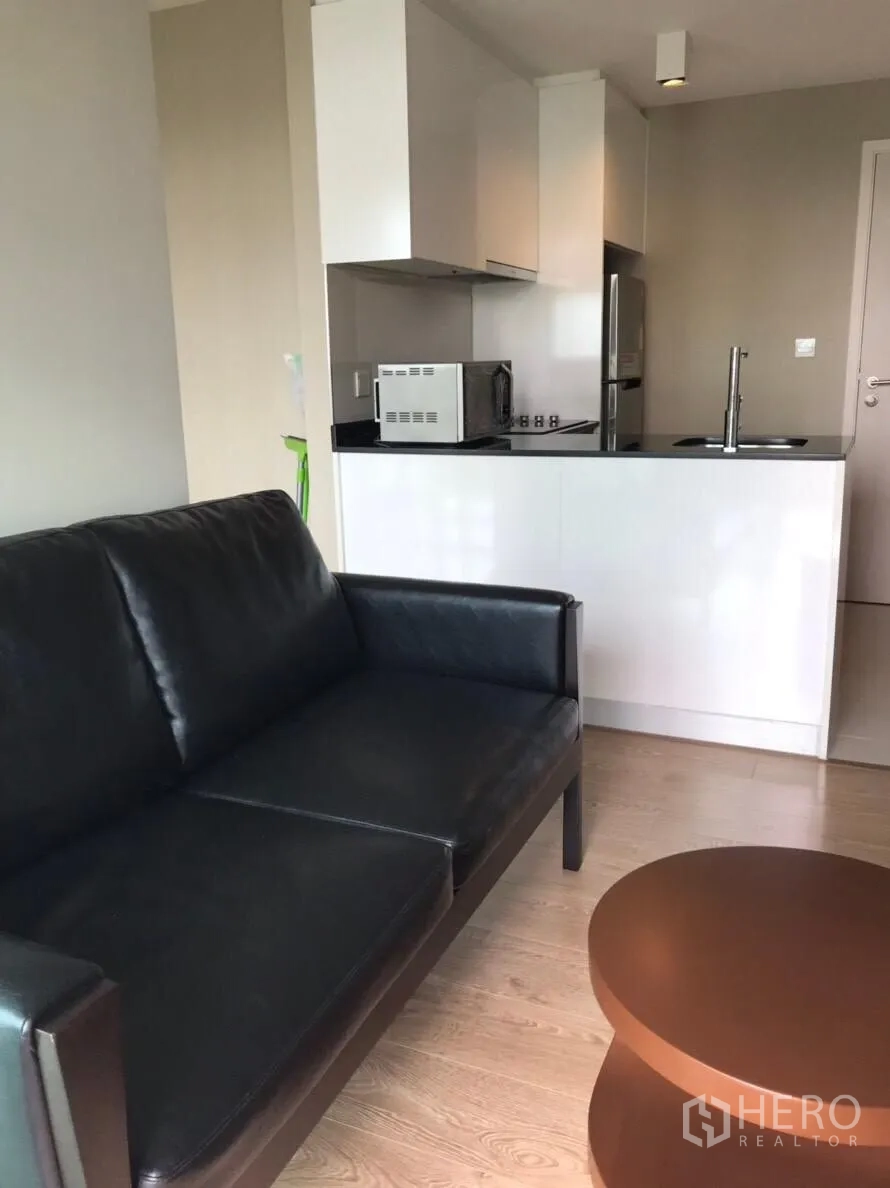 Condominium for rent in Watthana, Bangkok - Open-plan living area with black leather sofa facing a compact white kitchenette.