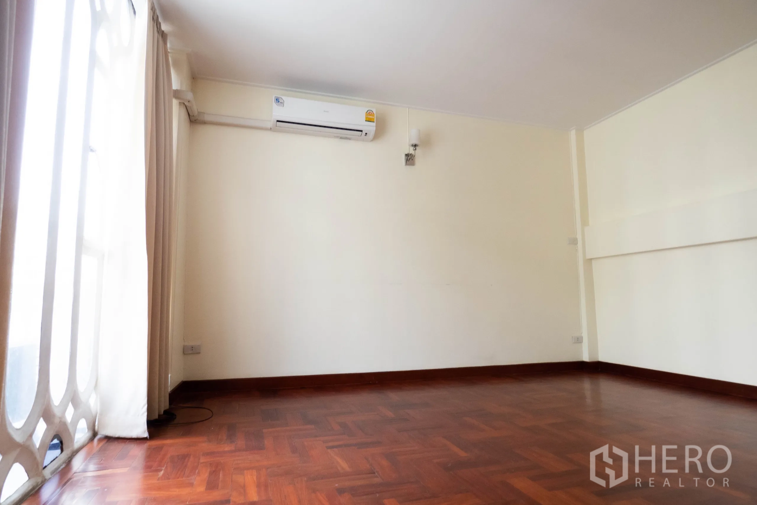 Townhouse for rent in Khlong Toei, Bangkok - Empty room with parquet floor, wall AC and tall arched window with curtains.
