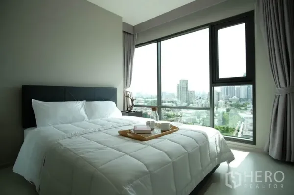 Condominium for rent in Khlong Toei, Bangkok - Bedroom with large windows, white bedding and panoramic skyline view.