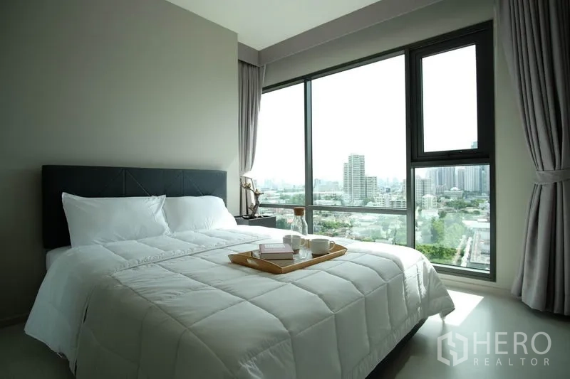 Condominium for rent in Khlong Toei, Bangkok - Bedroom with large windows, white bedding and panoramic skyline view.