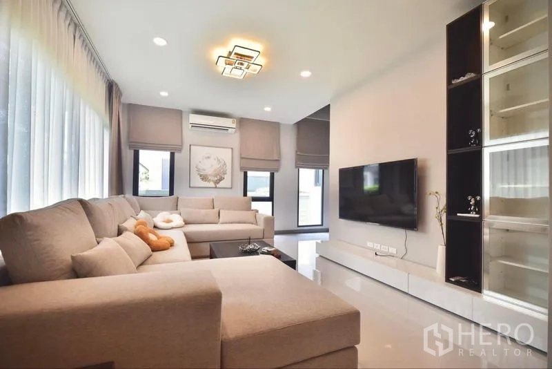Detached House for rent in Bang Phli, Samut Prakan - Living room with large sectional sofa, TV and floor-to-ceiling windows.
