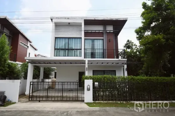 Detached House for rent in Prawet, Bangkok - Modern two-storey detached house in Prawet with gated carport, balcony, and hedge-lined frontage.
