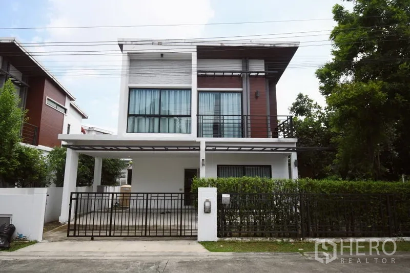 Detached House for rent in Prawet, Bangkok - Modern two-storey detached house in Prawet with gated carport, balcony, and hedge-lined frontage.