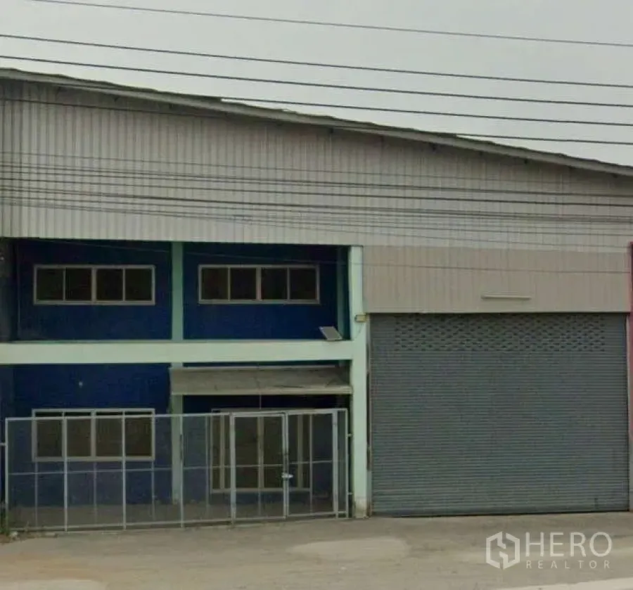 Warehouse for rent in Bang Bua Thong, Nonthaburi - Exterior of a steel-clad warehouse with high gable roof, wide roller-shutter door, two-storey blue office section and gated entrance in Bang Bua Thong.