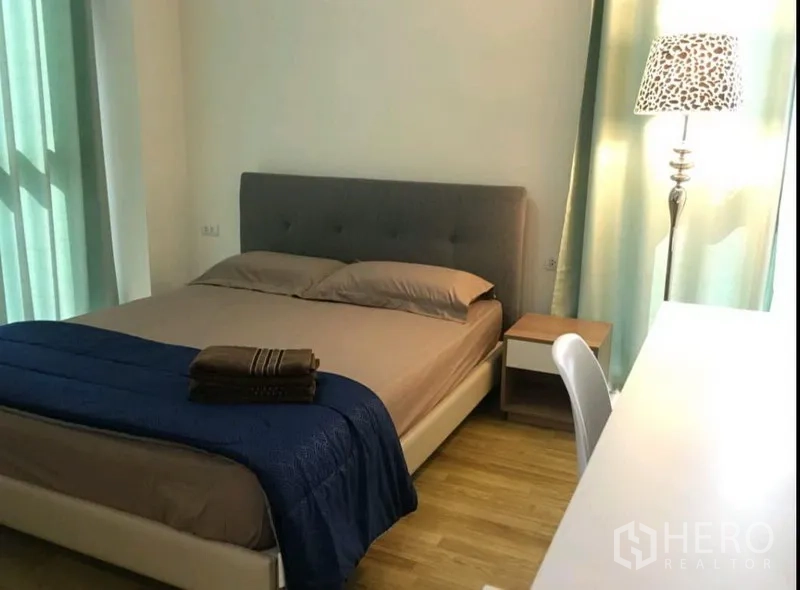 Condominium for rent in Phra Khanong, Bangkok - Master bedroom with upholstered double bed, bedside table, desk, floor lamp and soft green curtains over wood flooring.