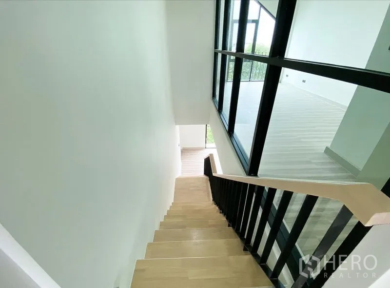 带办公室的家 出租或出售在Bang Na, Bangkok - 自楼梯向下的视角，浅木踏步与黑色扶手，旁为高窗墙。