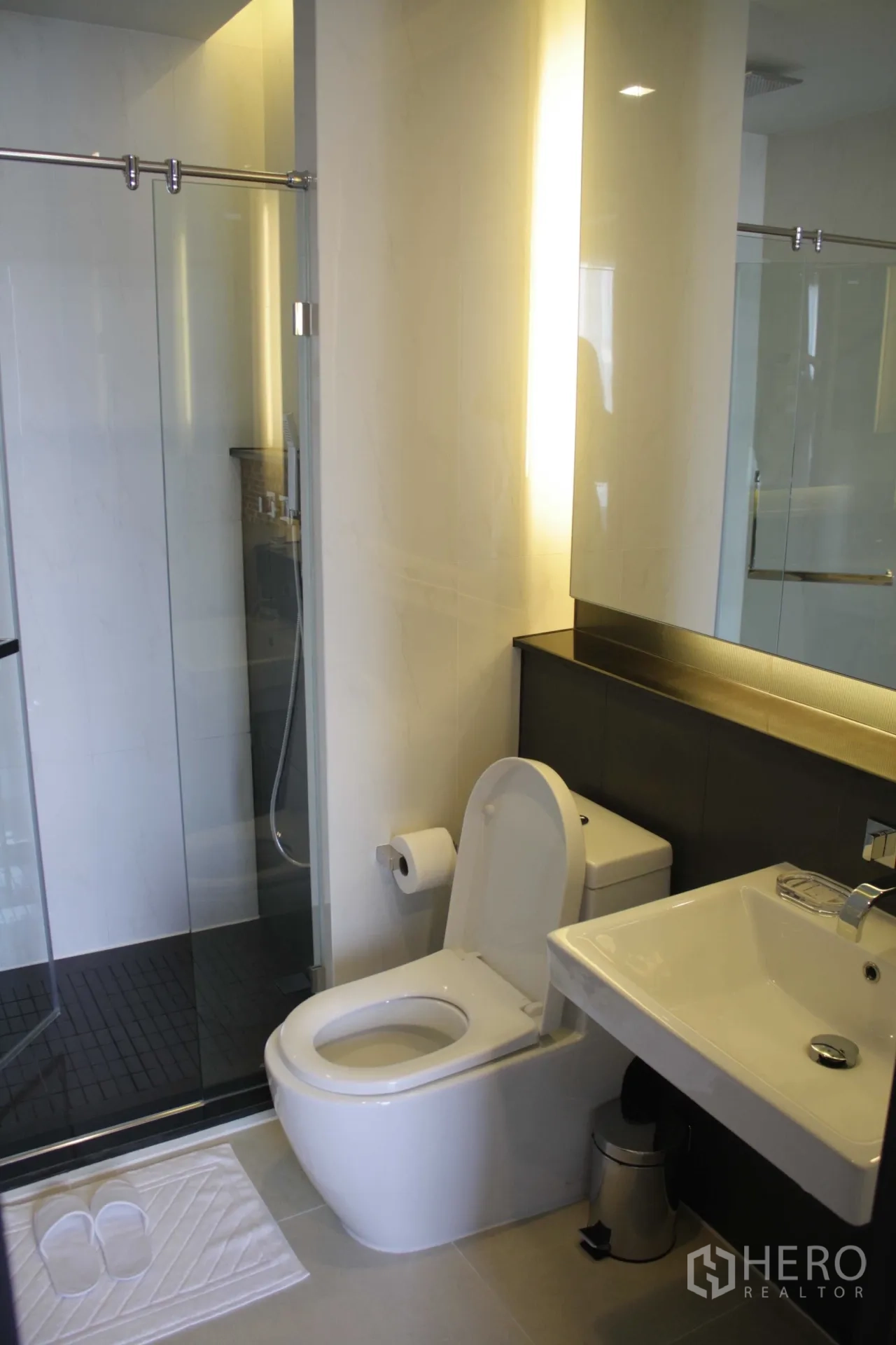 Condominium for rent in Huai Khwang, Bangkok - Modern bathroom with glass-enclosed shower, wall-mounted sink, and toilet under warm lighting.