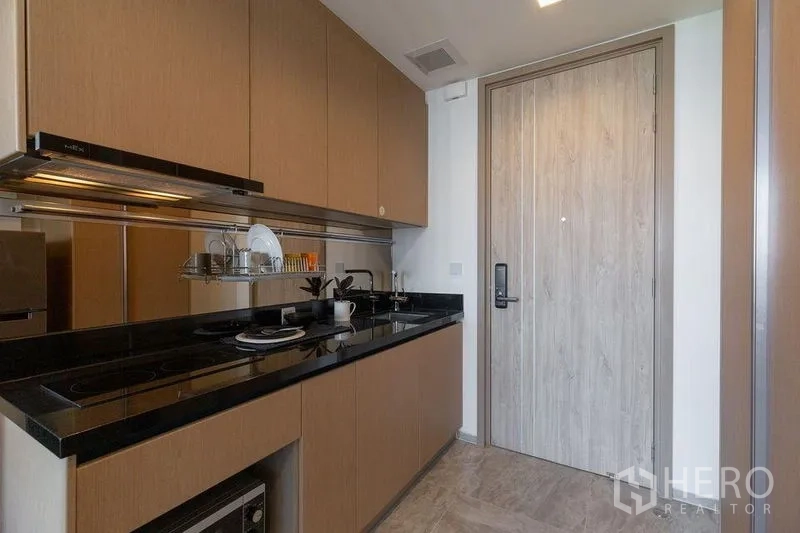 คอนโด ให้เช่าในWatthana, Bangkok - ครัวแบบแกลลี่ เคาน์เตอร์หินสีดำ เตาไฟฟ้า อ่างล้างจาน และตู้เก็บของเหนือศีรษะใกล้ประตูทางเข้า