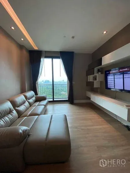 คอนโด ให้เช่าในBangkok - ห้องนั่งเล่นโซฟาหนัง ผนังติดทีวี และประตูกระจกสไลด์ออกระเบียงรับวิวสวนและเมือง