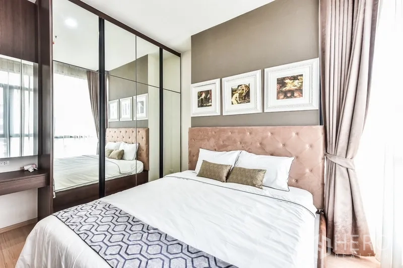 Condominium for rent or sale in Huai Khwang, Bangkok - Master bedroom with padded headboard and full-height mirrored closets beside a window.