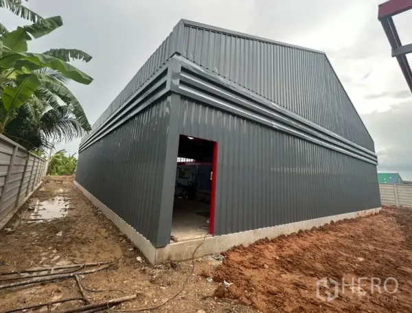 货栈 出租在Lam Luk Ka, Pathum Thani - 仓库转角近景，灰色波纹金属墙体、红色门框入口，以及沿围墙的通道。