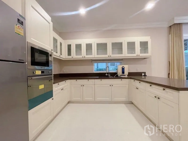 Detached House for rent or sale in Bang Phli, Samut Prakan - U‑shaped kitchen with white shaker cabinets, glass uppers, built‑in oven, and long counters.