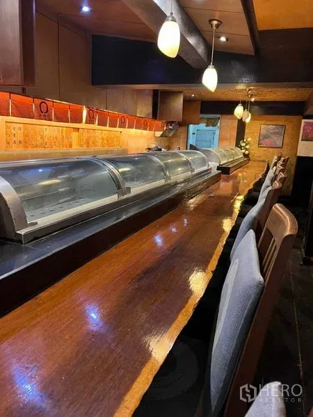 อาคารพาณิชย์ ให้เช่าในKhlong Toei, Bangkok - เคาน์เตอร์ซูชิไม้พร้อมตู้แช่และเก้าอี้สูงในบรรยากาศไฟส้มอุ่นภายในอาคารพาณิชย์