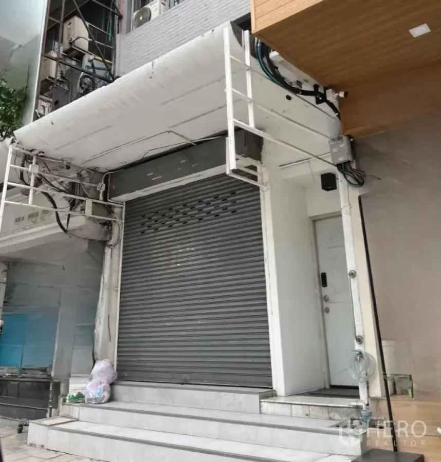 零售空间 出租在Watthana, Bangkok - 一层店面外观，卷闸门关闭，白色立面与台阶，临阿索克繁华街道。