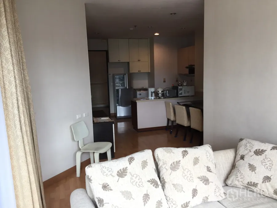 Condominium for rent in Khlong Toei, Bangkok - Open-plan lounge leading to kitchen with dining chairs and tall curtains.