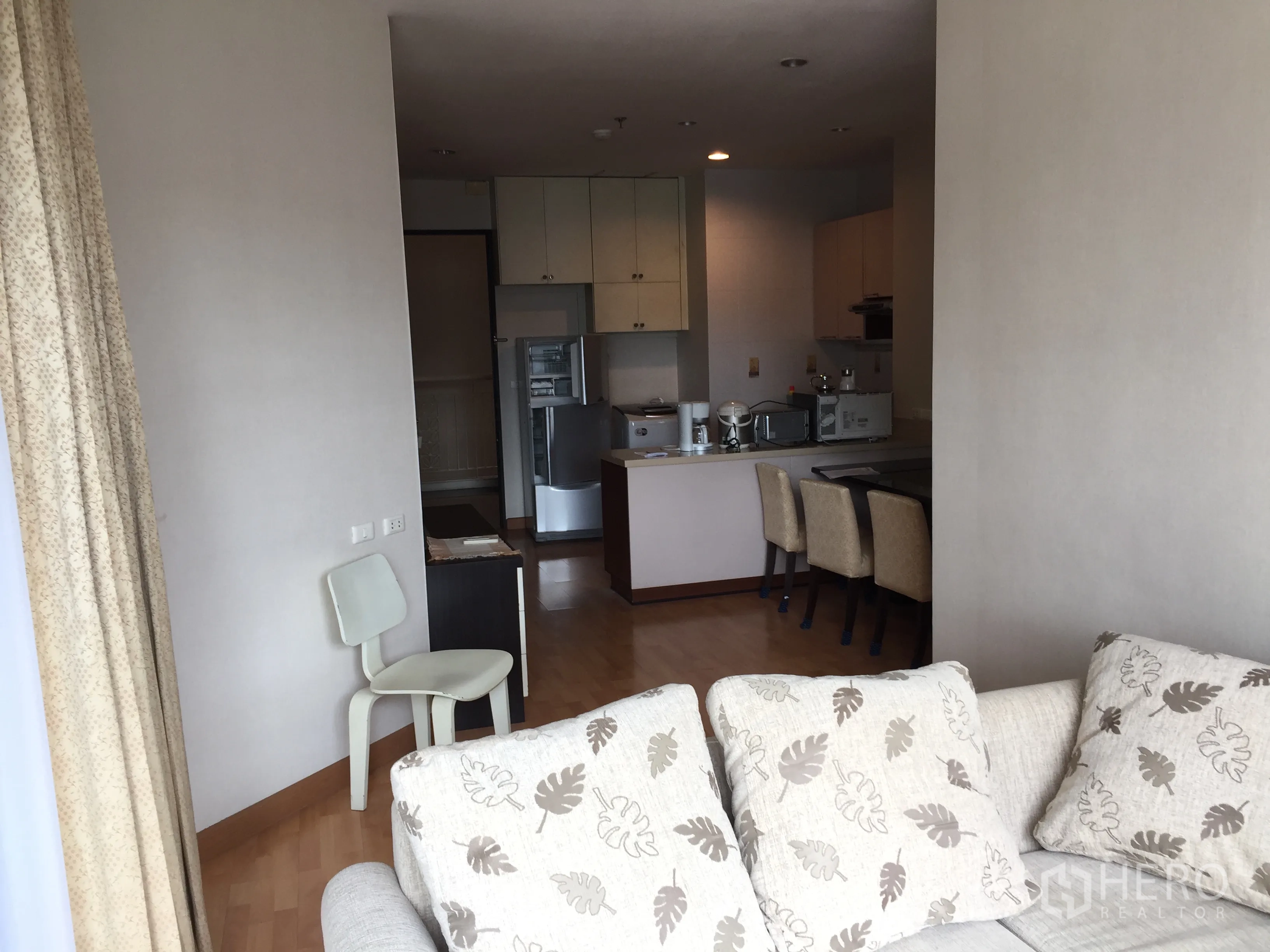 Condominium for rent in Khlong Toei, Bangkok - Open-plan lounge leading to kitchen with dining chairs and tall curtains.