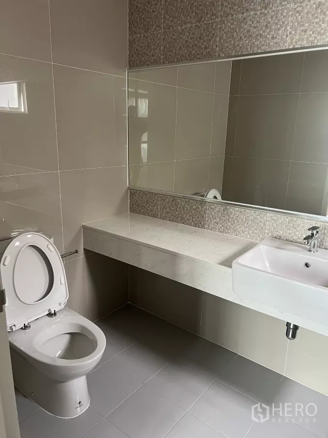 Detached House for rent or sale in Don Mueang, Bangkok - Bathroom featuring long vanity counter, large mirror and toilet on grey tiles.